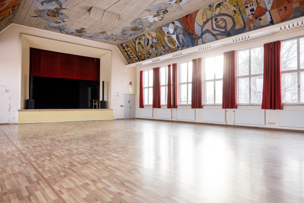 Evenemangslokal Albrektssalen: Allrum med scen, höga fönster, röda gardiner och målning i taket.