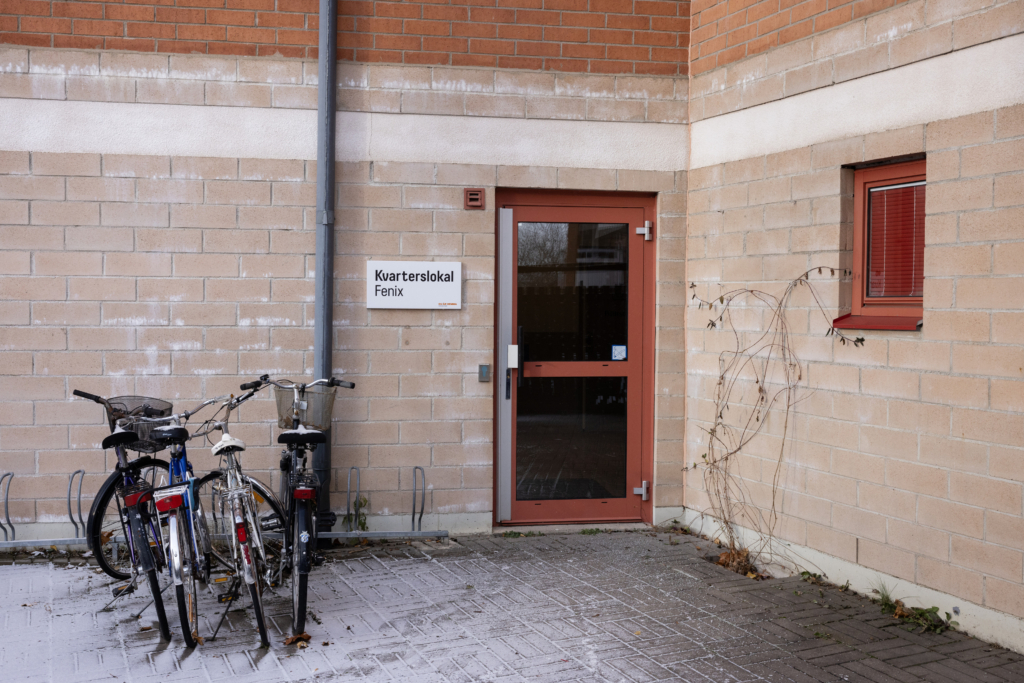 Kvarterslokal Fenix: Tegelfasad med entrédörr till kvarterslokalen. Cykelställ finns på vänster sida om dörren.