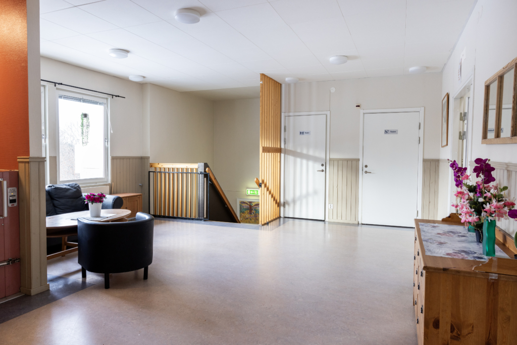 Kvarterslokal Mässen: En tvåsitssoffa och fåtölj med soffbord i korridorsutrymmet. En RWC, WC och trappa samt hiss till nedervåning.