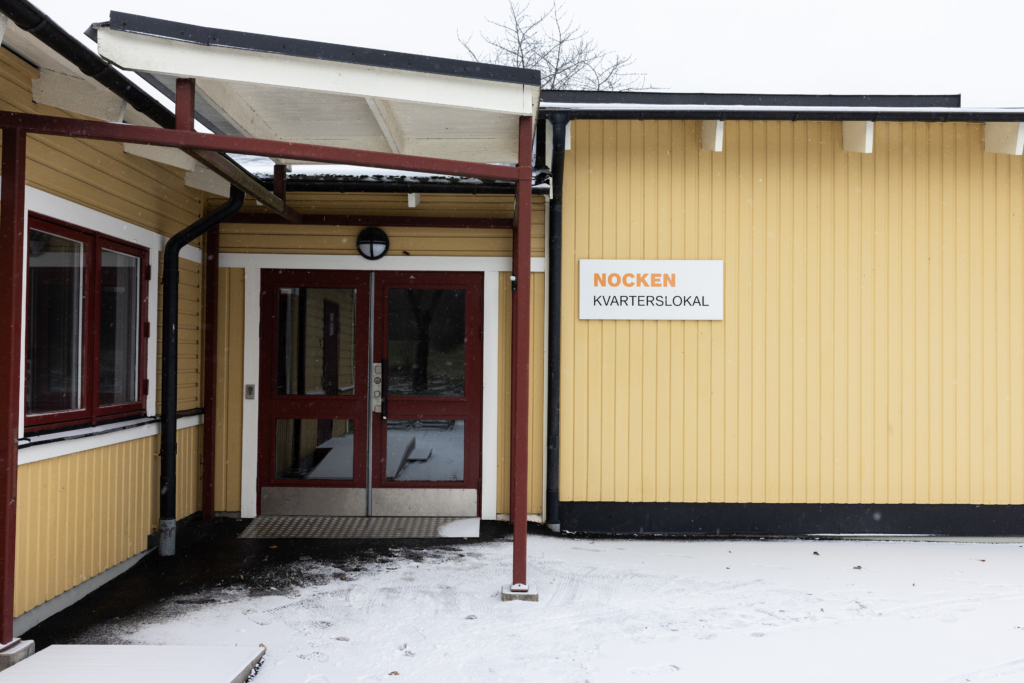 Kvarterslokal Nocken: En gul byggnad med en röd dörr och en skylt.