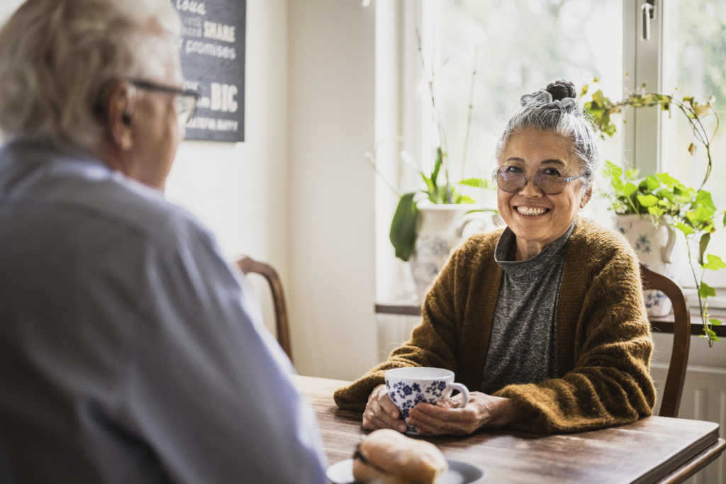 Ett äldre par sitter i ett kök och fikar. Kvinnan ler mot kameran. 