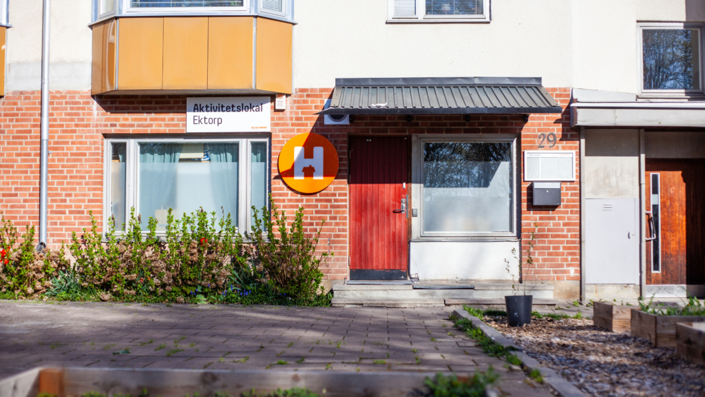 Fasadbild på entrén till Aktivitetslokalen. På vänster sida om den röda dörren sitter en skylt. På höger sida är entrén till lägenhetshuset som Aktivitetslokalen ligger i.