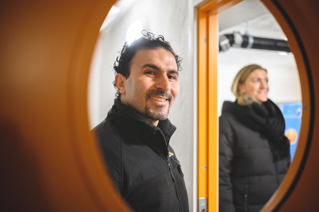 En man tittar in genom ett fönster i en dörr och ler. En suddig kvinna i bakgrunden. 