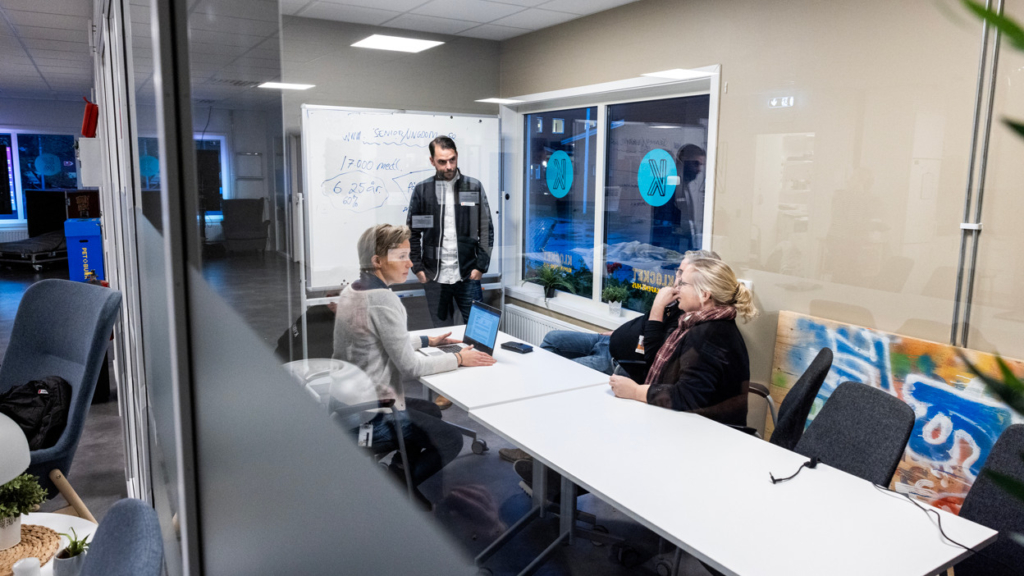 Klocket, Kunskapens hus. Tre personer sitter runt ett konferensbord och lyssnar till en person som står vid en whiteboard-tavla.