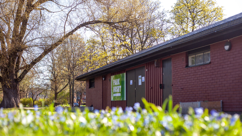 Fasaden på Parkhuset, en röd tegelbyggnad med svarta dörrar. Blommor i förgrunden och en stor ek i bakgrunden.