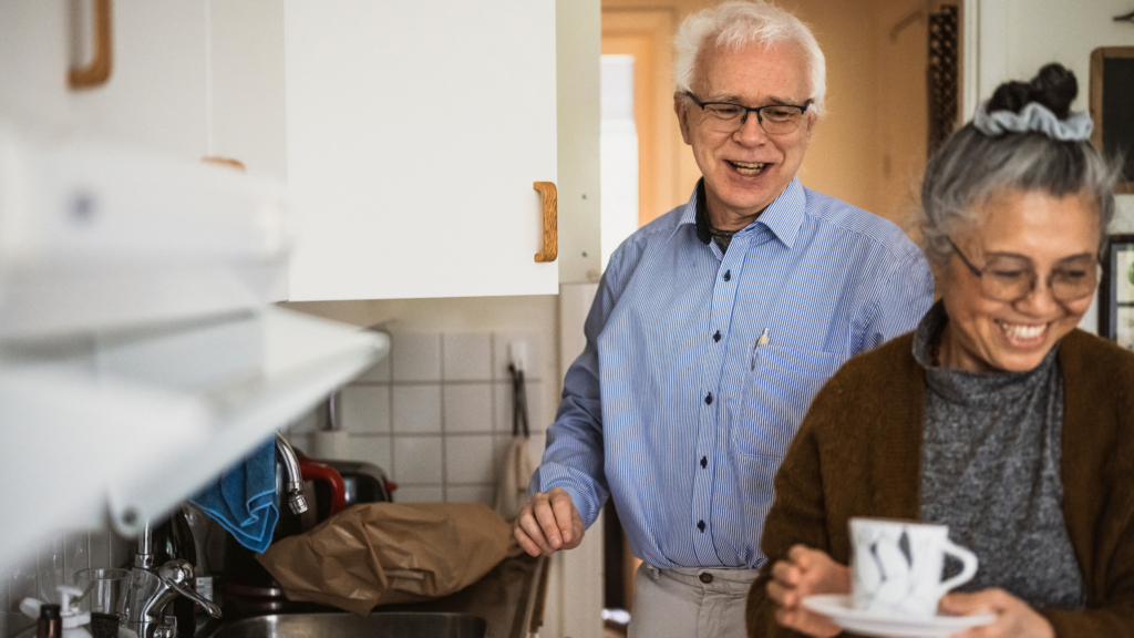 Ett äldre par står vid en köksbänk. Båda kollar bort från kameran.