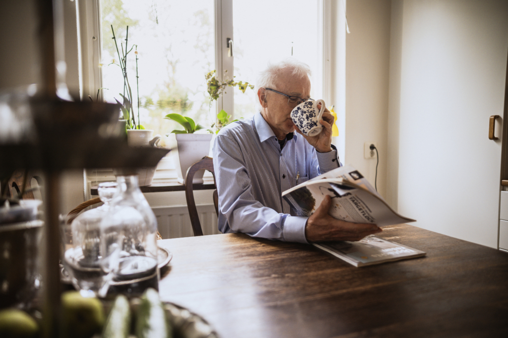 En senior man som dricker kaffe och läser tidningen vid köksbordet.