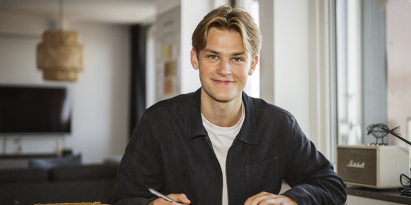 En ung kille sitter vid ett bord och skriver i en anteckningsbok. Han tittar in i kameran och ler.