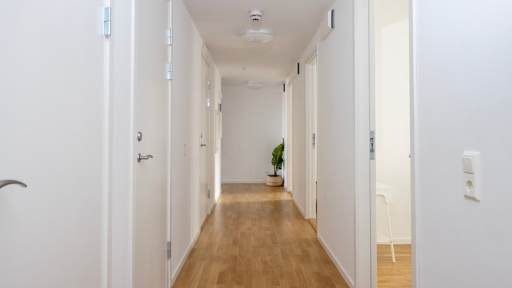 Bo tillsammans-lägenhet: En lång korridor med vita väggar och trägolv. Längst med högra väggen syns tre dörrar in till privata rum. Längst med vänstra väggen finns tre dörrar till WC och duschrum.