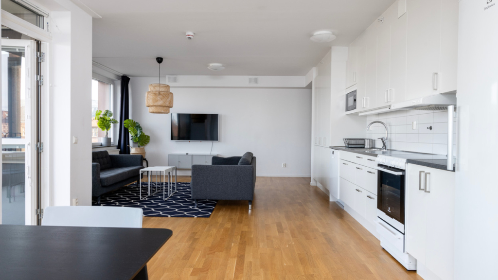 Öppen planlösning i en Co-living-lägenhet med kök, köksbord och två soffor som står runt ett soffbord. Tv hänger på väggen och en tv-bänk står under.
