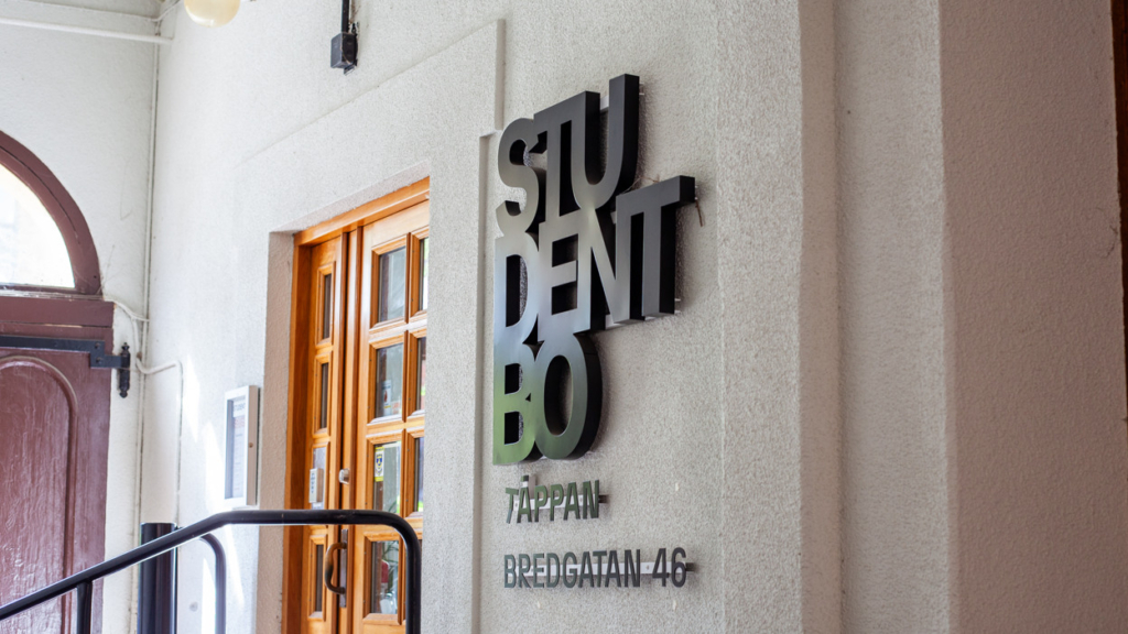 Entrén till Studentbos huvudkontor som ligger i ett valv med stor mörkröd port. Entrédörren är i massivt trä och till höger om dörren finns en Studentbo-skylt.