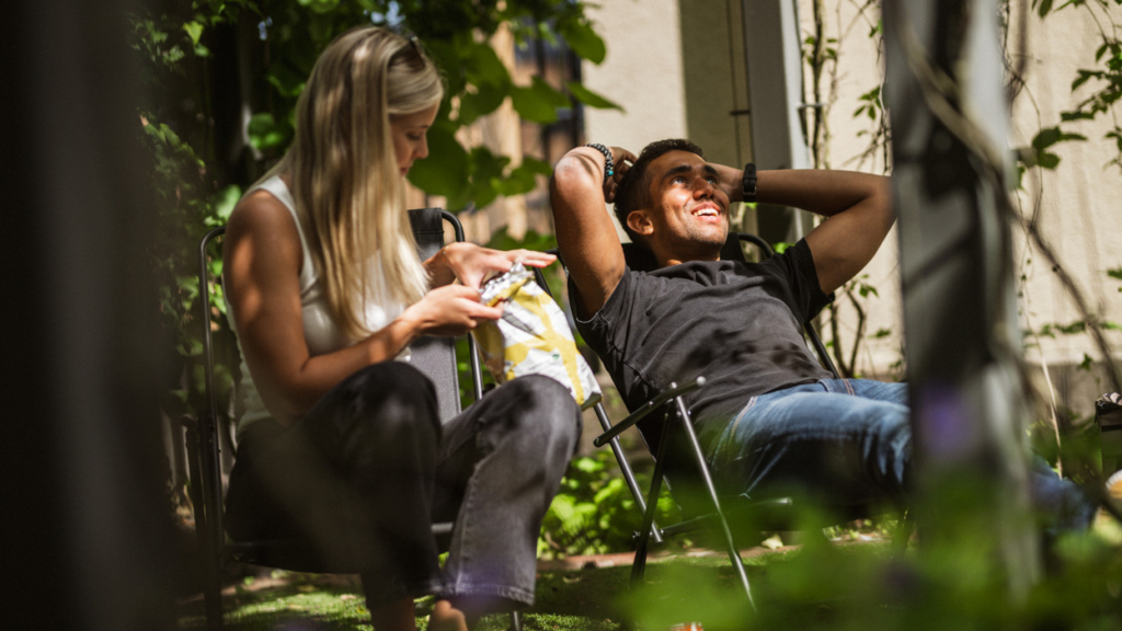 Två studenter som sitter i solstolar under ett grönskande träd.